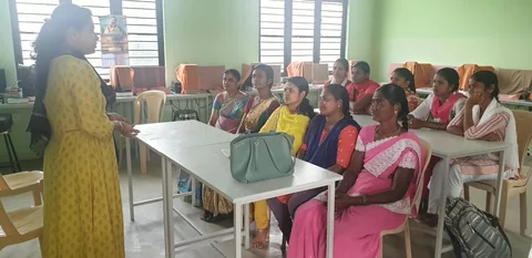 Women seated around classroom tables, listening to a speaker beside them.